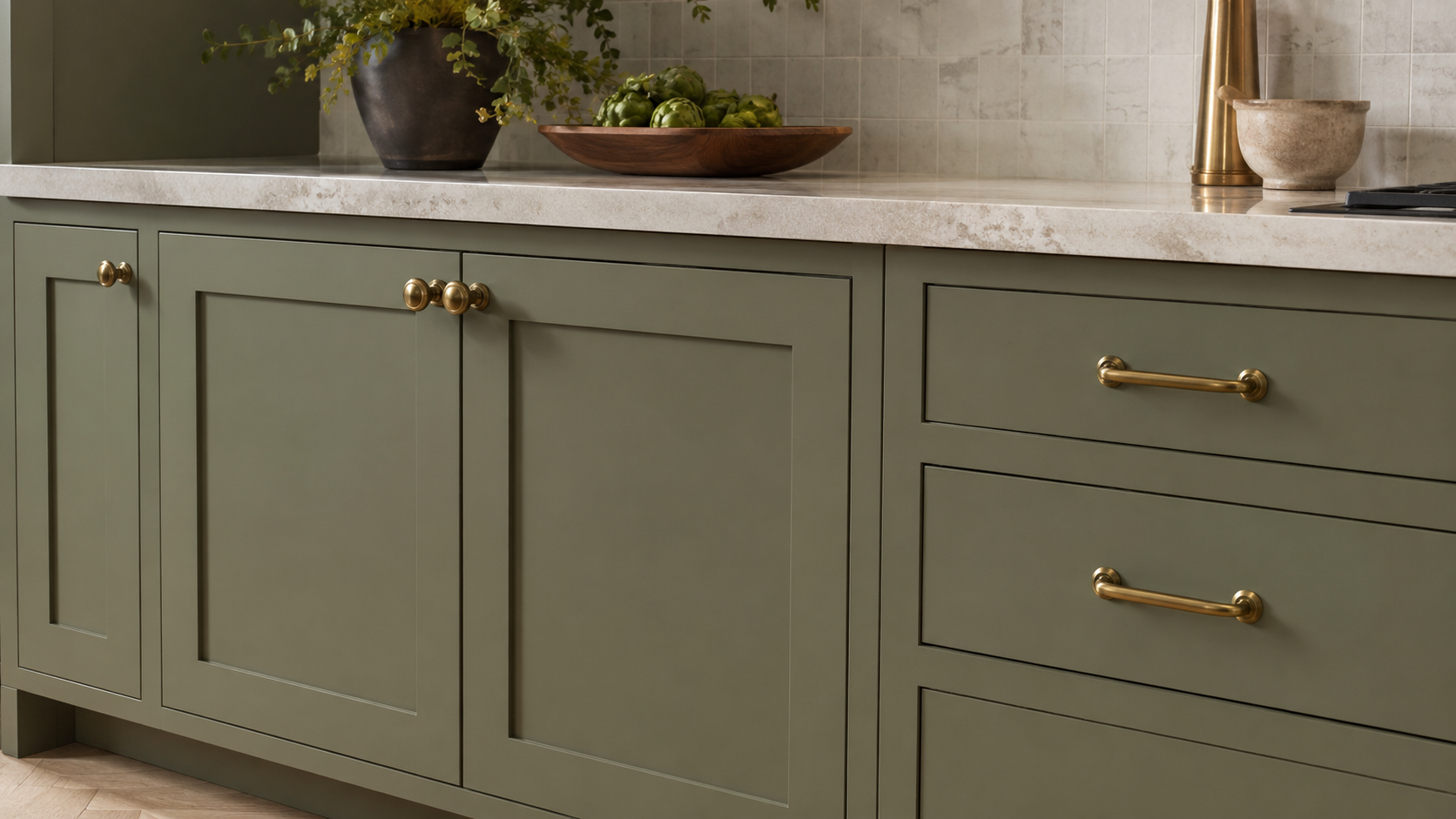 Close-up of muted sage or olive green kitchen cabinets with elegant brass knobs and pulls, pale stone counter, and refined modern detail