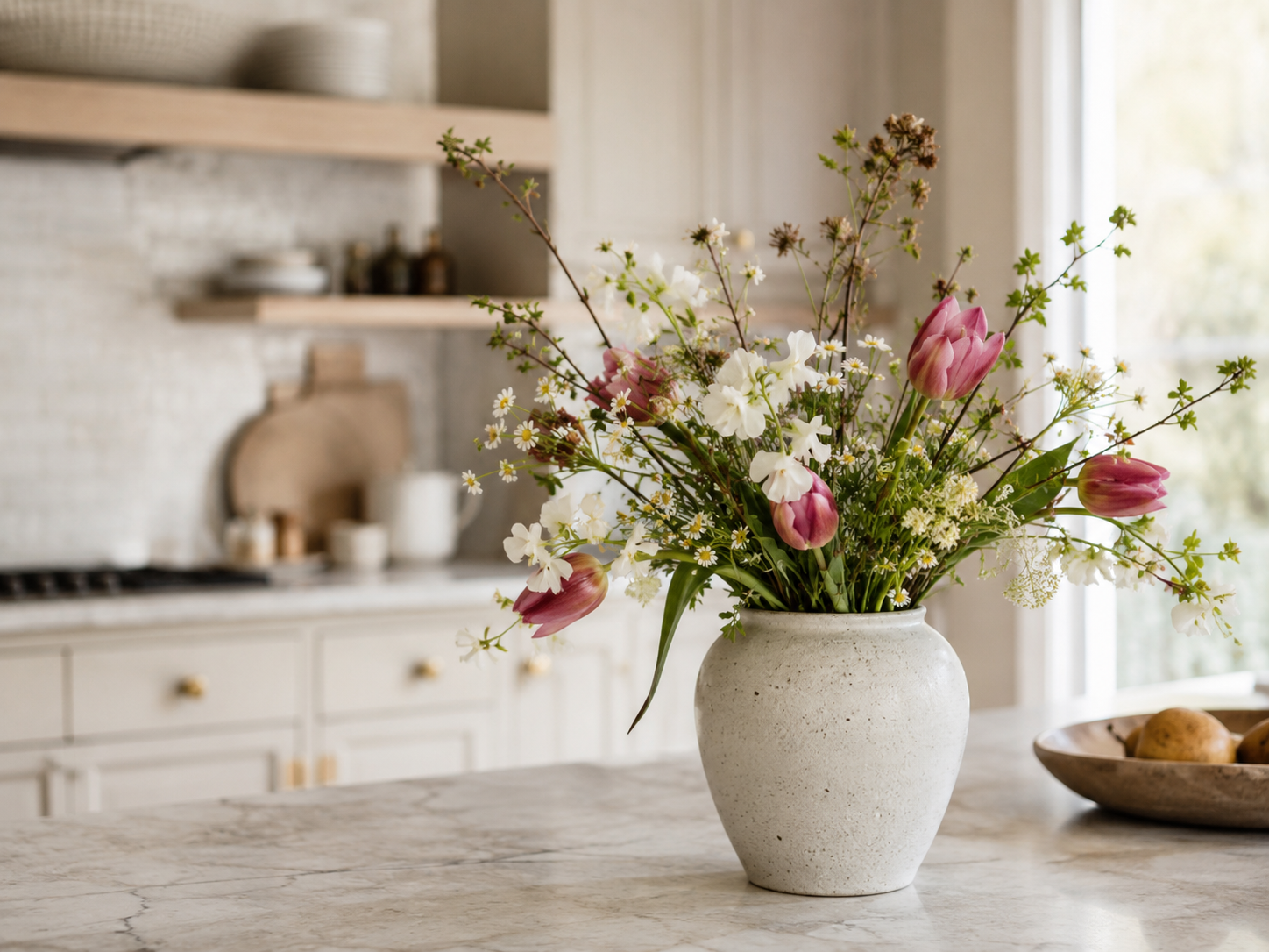 Kitchen counter or island with ceramic vase of seasonal flowers, greenery, light stone countertop, neutral cabinets, and soft daylight
