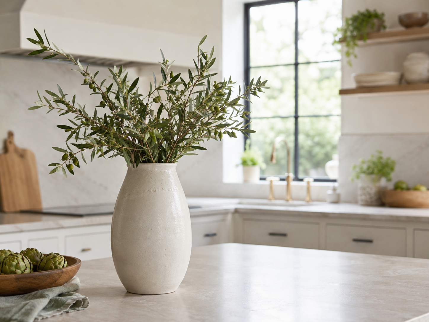 Kitchen counter with tall off-white ceramic vase filled with olive branches or eucalyptus, pale stone countertop, cream cabinetry, and daylight