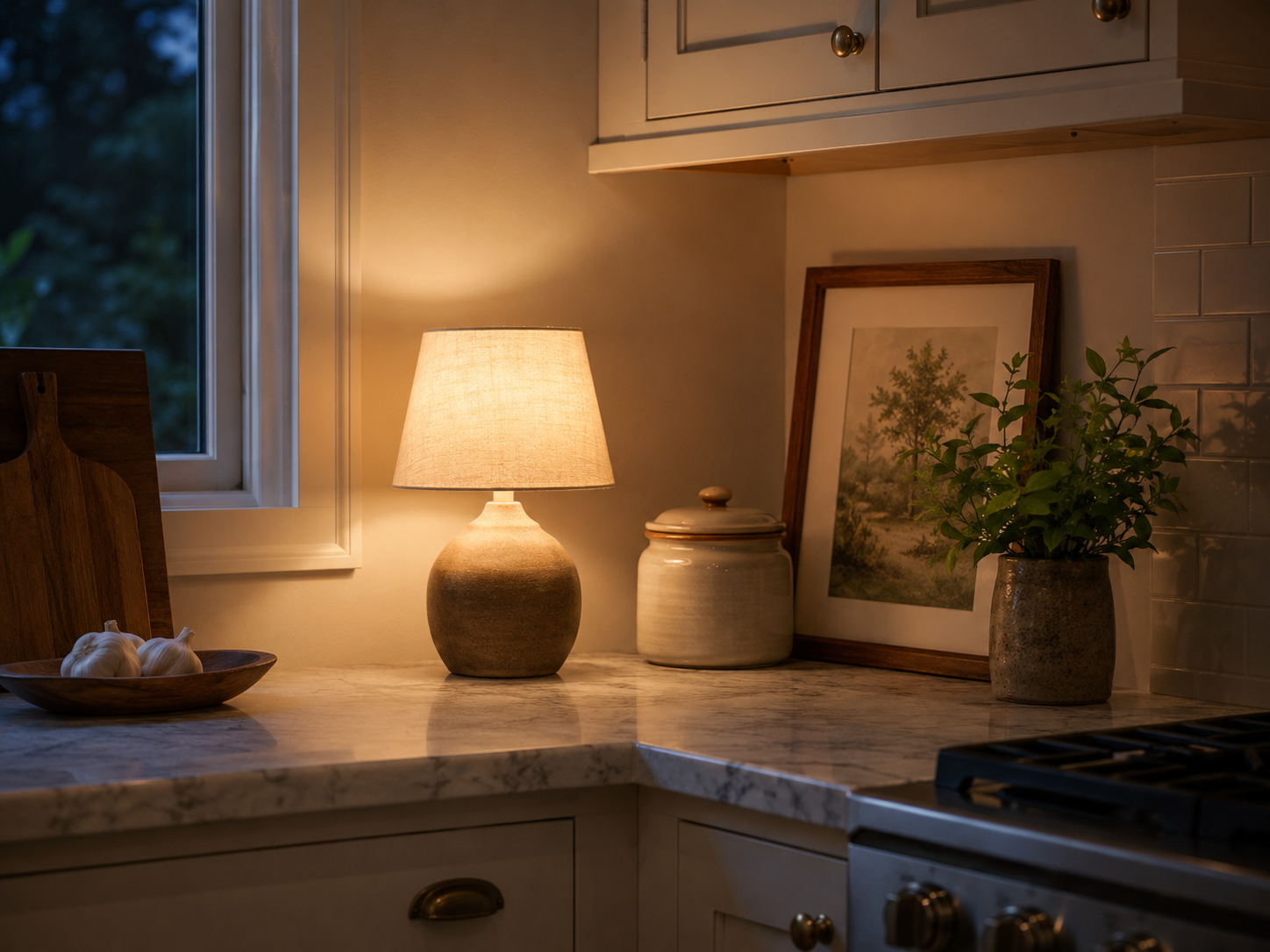 Evening kitchen counter corner with small warm lamp beside ceramic jar, framed print, plant, muted cream cabinets, and soft shadows