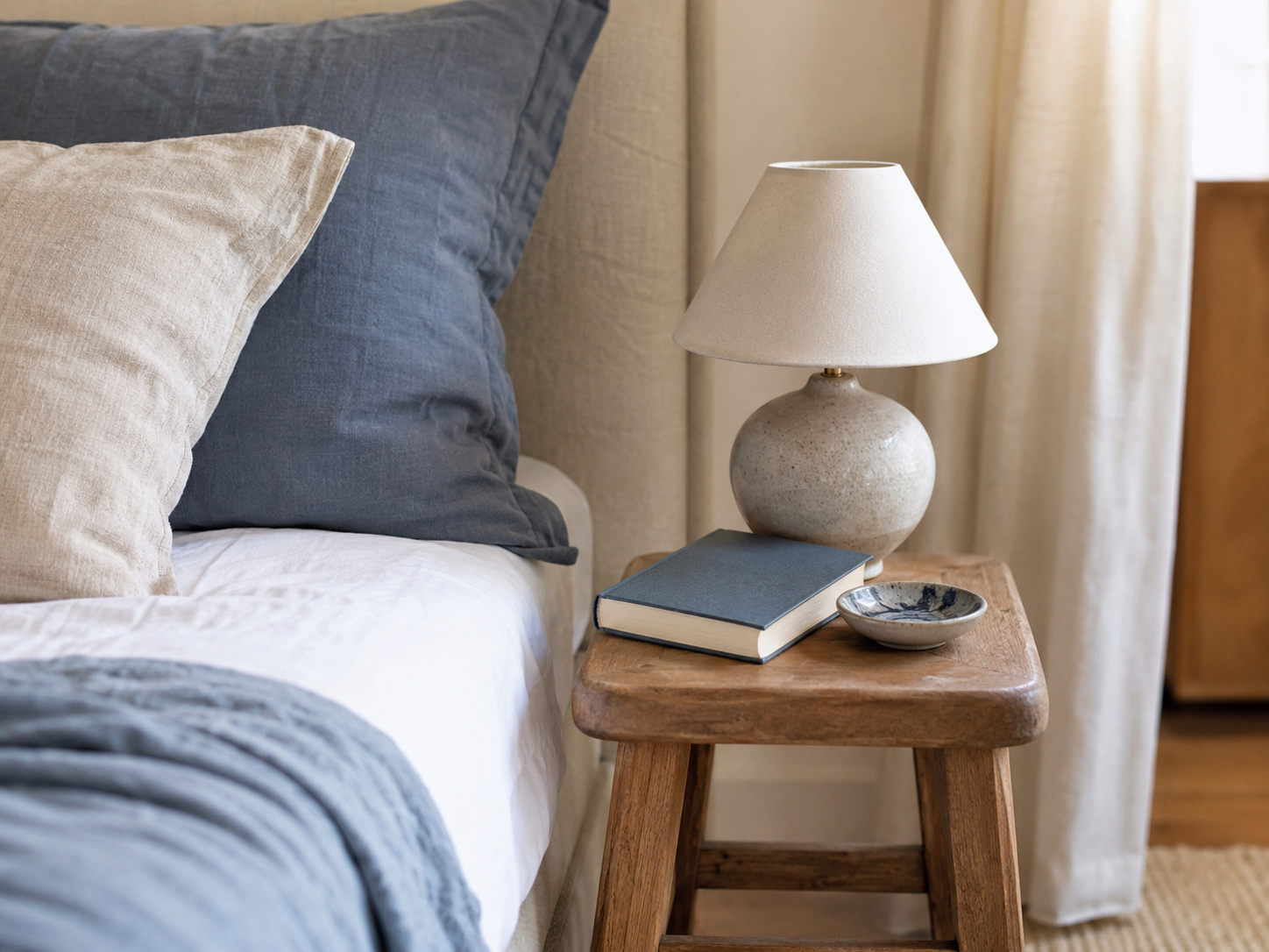 Close-up of compact apartment bedroom nightstand or stool with lamp, book, ceramic dish, soft bedding, and light curtain