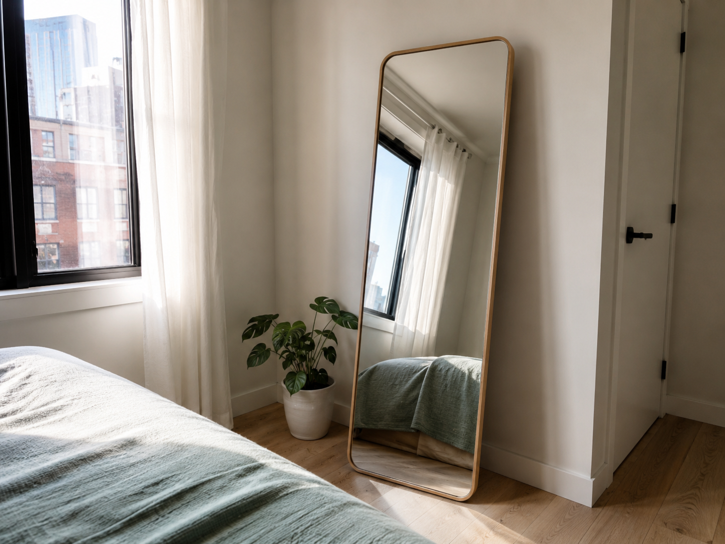 Apartment bedroom with a tall leaning mirror near a window, compact bed, light curtains, plant, and pale oak floor