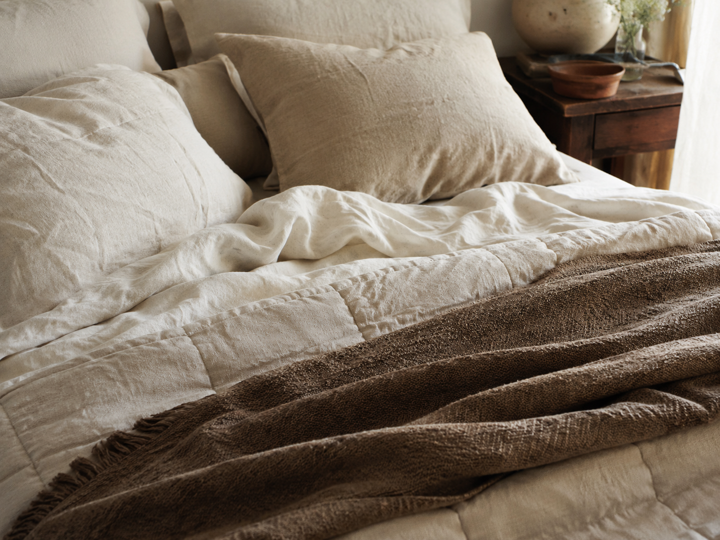 Close-up of layered linen bedding with rumpled cream sheets, beige pillows, quilt, and taupe throw blanket