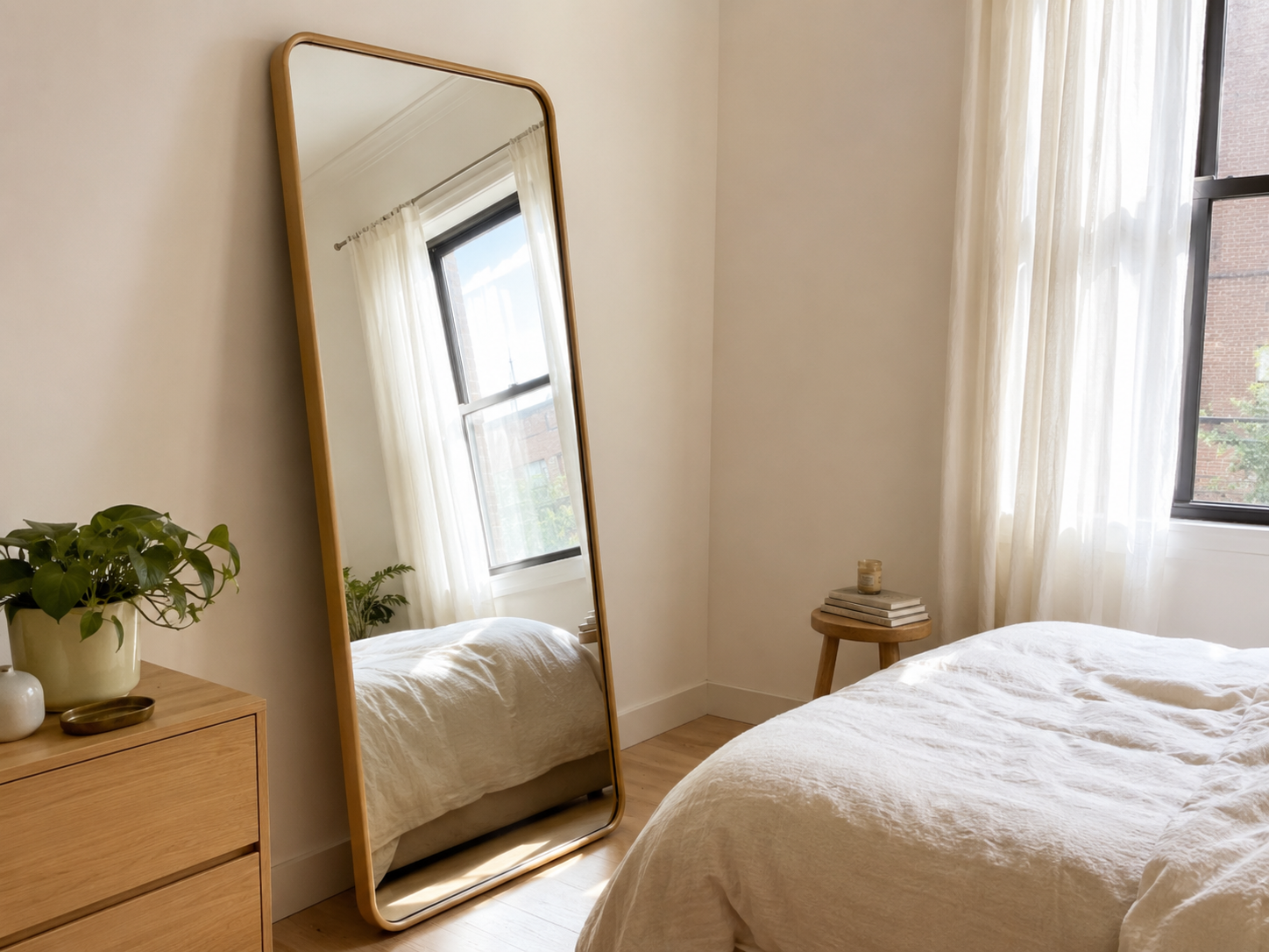 Small bedroom with a tall leaning mirror near a window reflecting daylight, compact bed, curtains, plant, dresser or stool, and light wood floor