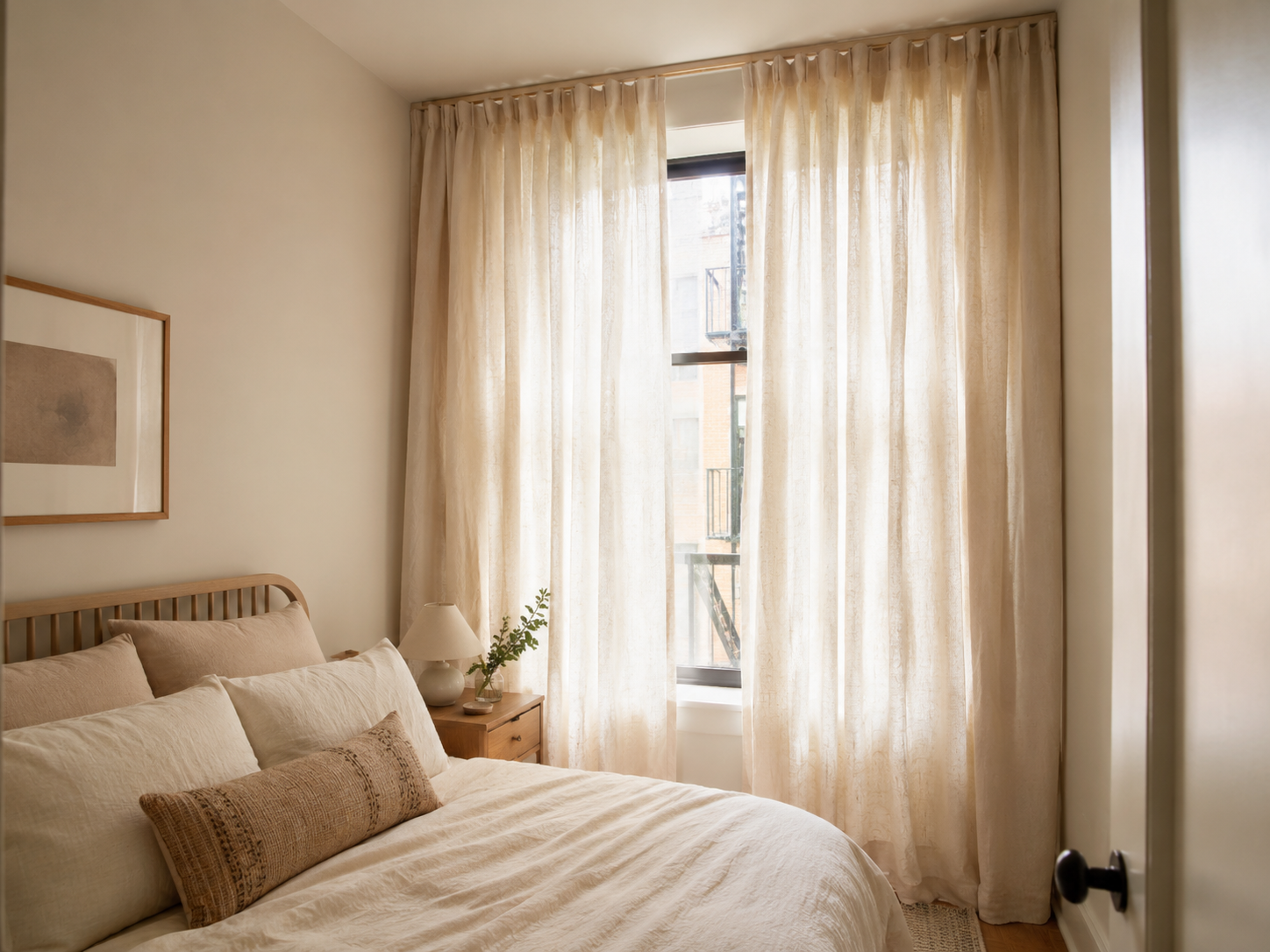 Small bedroom window with tall cream linen curtains, soft daylight, neutral bedding, and narrow nightstand